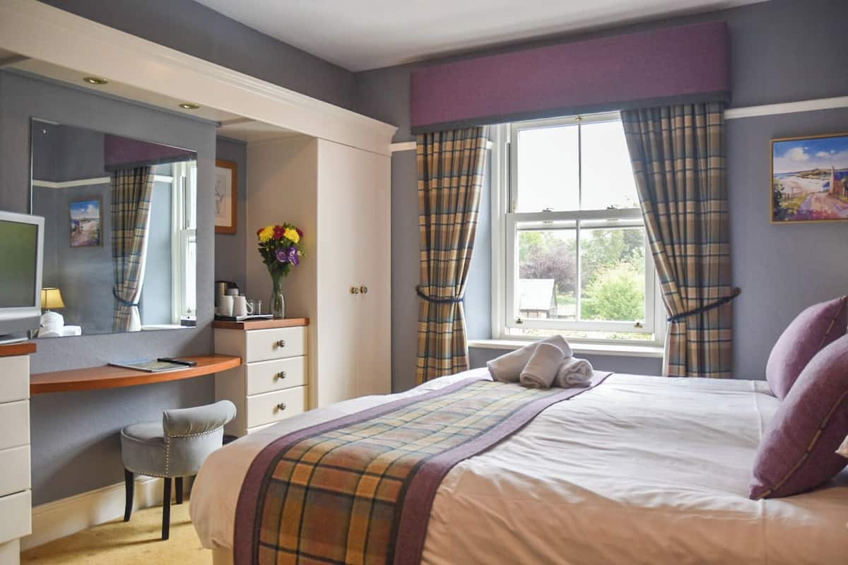 A bedroom with a bed, a desk, a chair, a TV, and a window with curtains.