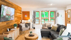 A modern living room with a wooden wall, a flat-screen TV, a sofa, a coffee table, and a view of greenery through large windows. - Perfect Summer Getaway-Near Storyland (Bartlett)
