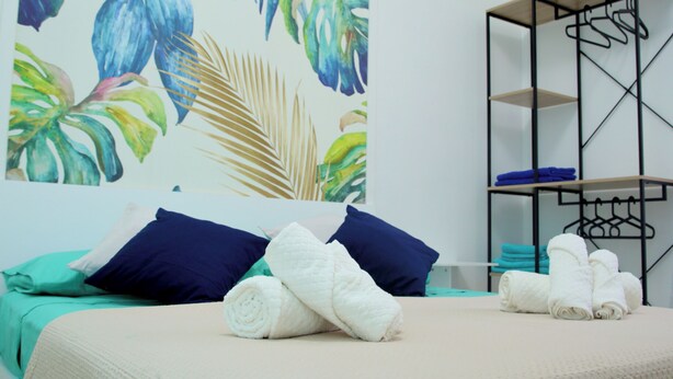 A neatly made bed with blue and white pillows, a wooden shelf unit with folded towels, and a tropical leaf-patterned wall. - DOMUS GREEN 20 minutes from the Vatican and the Historic Center of Rome (Roma)