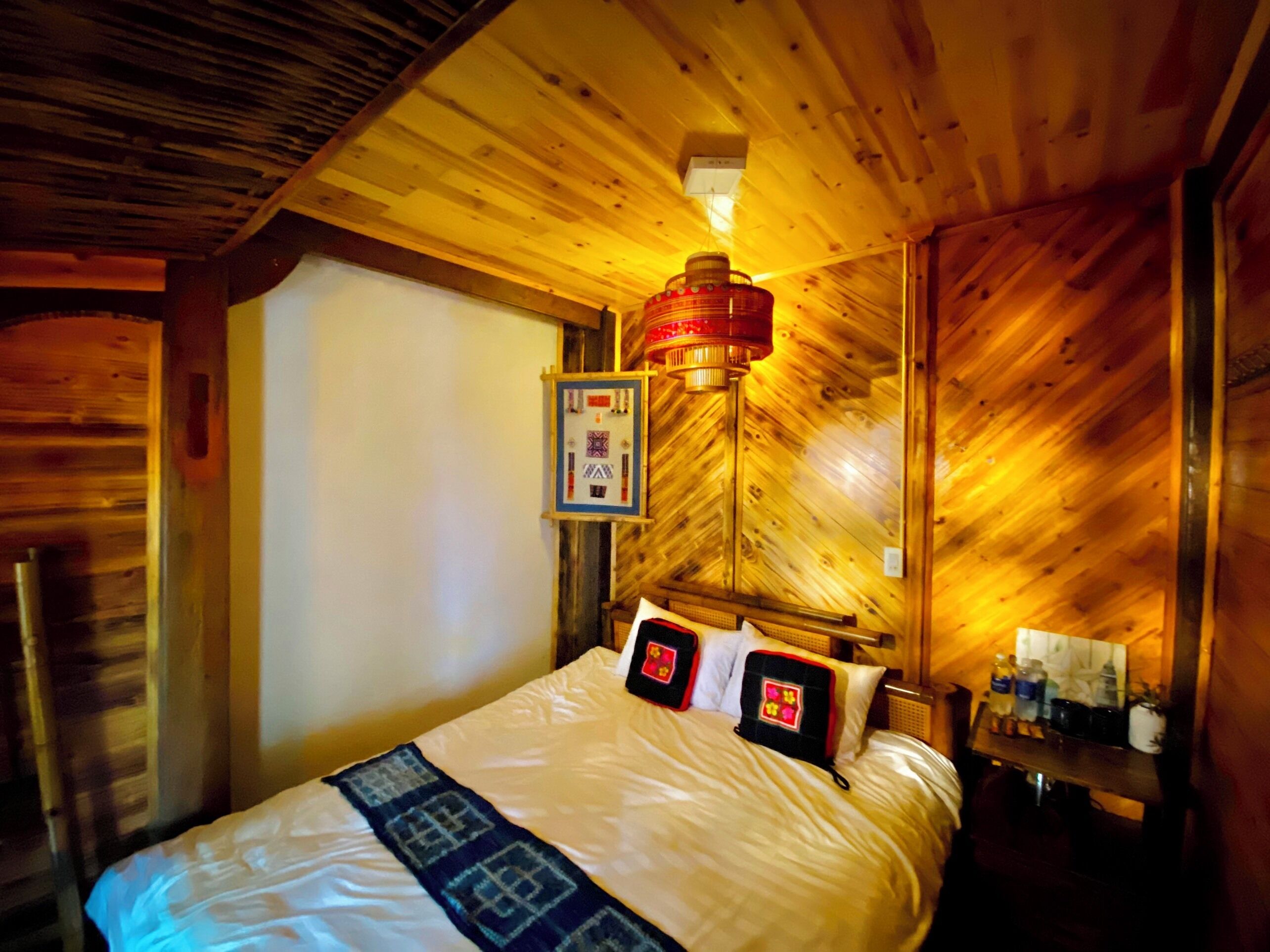 A wooden cabin room with a bed, a wall-mounted lamp, and a small table with bottles.