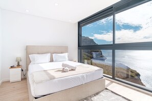 Une chambre à coucher moderne avec une grande fenêtre donnant sur la côte, un lit soigneusement fait et une table de chevet avec une lampe.