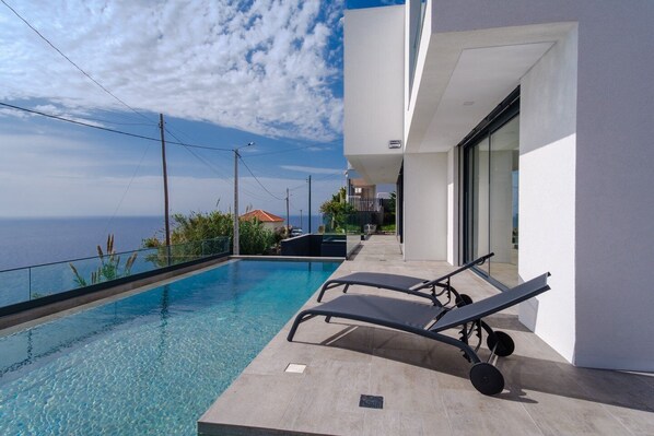 Une piscine extérieure moderne, dotée de chaises longues, offrant une vue dégagée sur la mer et bordée d’un bâtiment blanc aux grandes fenêtres.