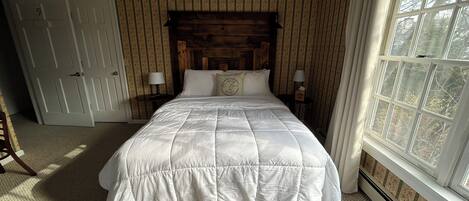 A bedroom with a large bed, two bedside tables with lamps, a wooden headboard, a door, and a window with a view of trees.