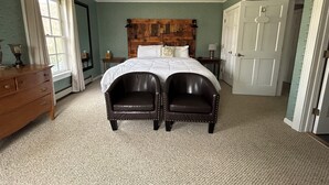 Une chambre à coucher avec une tête de lit en bois, un grand lit, deux fauteuils, une commode, une fenêtre avec des rideaux et une porte.