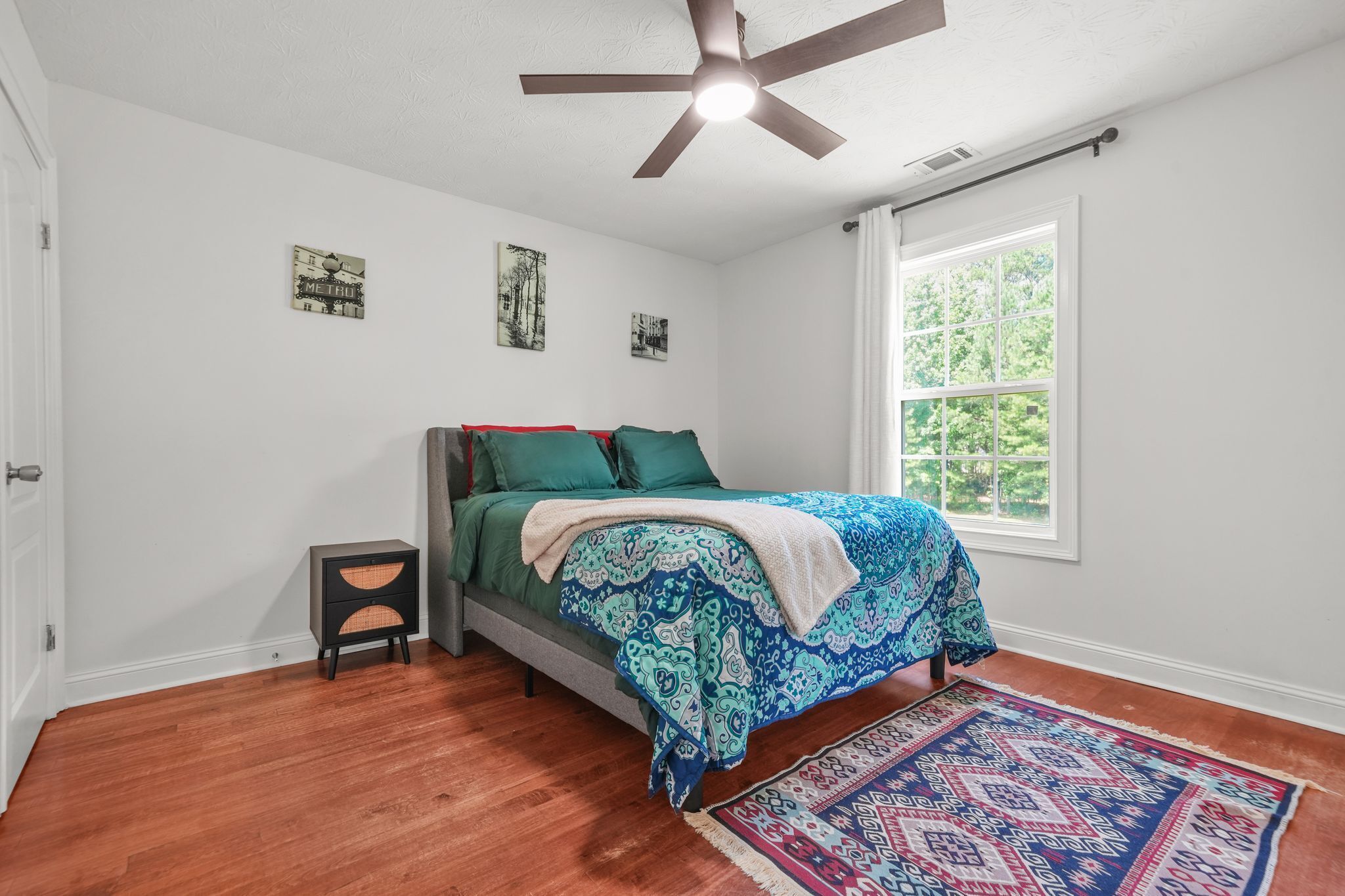 A bedroom with a bed, a ceiling fan, a window, and a small nightstand.