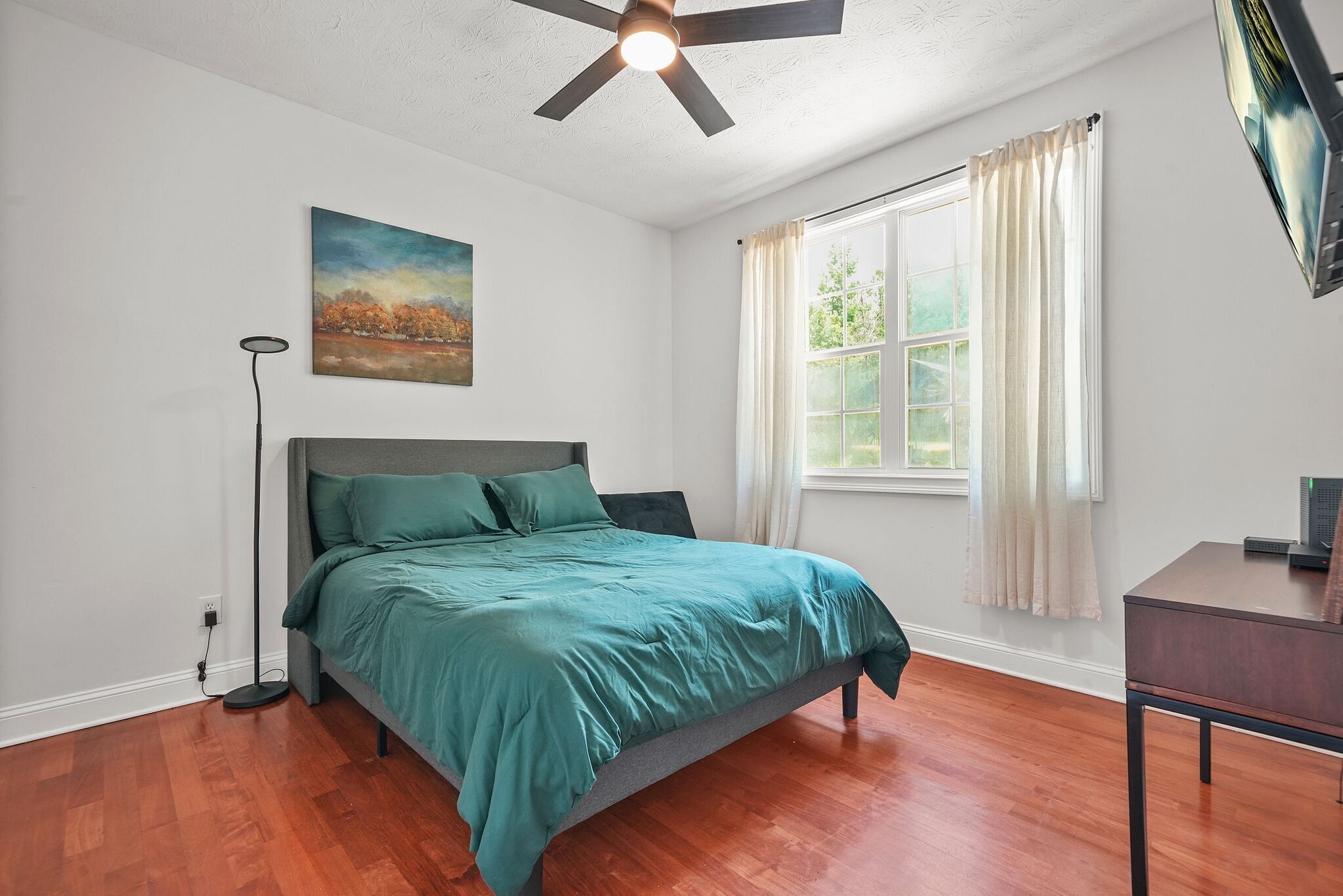 A bedroom with a bed, a ceiling fan, a window with curtains, a desk, and a painting on the wall.