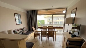 A compact hotel room with a kitchenette, a dining area with a table and chairs, a sofa, and a large window offering a view of the building and greenery outside.