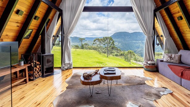 Ein gemütliches Zimmer mit Holzbalkendecke, einem großen Fenster mit Vorhängen, einem Sofa, einem Couchtisch, einem Fernseher und einem Kamin.