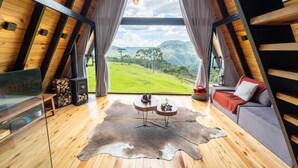 Ein gemütlicher Raum mit hohen Decken und Holzvertäfelung, ein großes Fenster mit Blick auf eine grüne Landschaft, eine Couch, ein Couchtisch und ein Kamin.