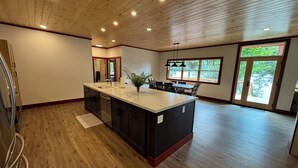 A spacious kitchen with a wooden ceiling, a large island with a sink, and a dining area with a view of the outdoors. - The Point House - Exclusive Lake Placid Boat-Access Home (Lake Placid)