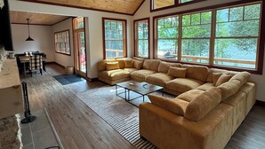 A spacious living area with a large sectional sofa, a ceiling fan, and large windows offering a view of the outdoors. - The Point House - Exclusive Lake Placid Boat-Access Home (Lake Placid)