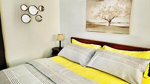 Un lit avec une couvre-lit à rayures jaunes et grises, deux oreillers et une tête de lit. Un tableau représentant un arbre est accroché au mur, et une petite table avec une lampe se trouve à proximité.