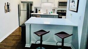 Une cuisine moderne avec des meubles blancs, un réfrigérateur en acier inoxydable, un four à micro-ondes et un évier. On trouve deux tabourets de bar sous l’îlot central.