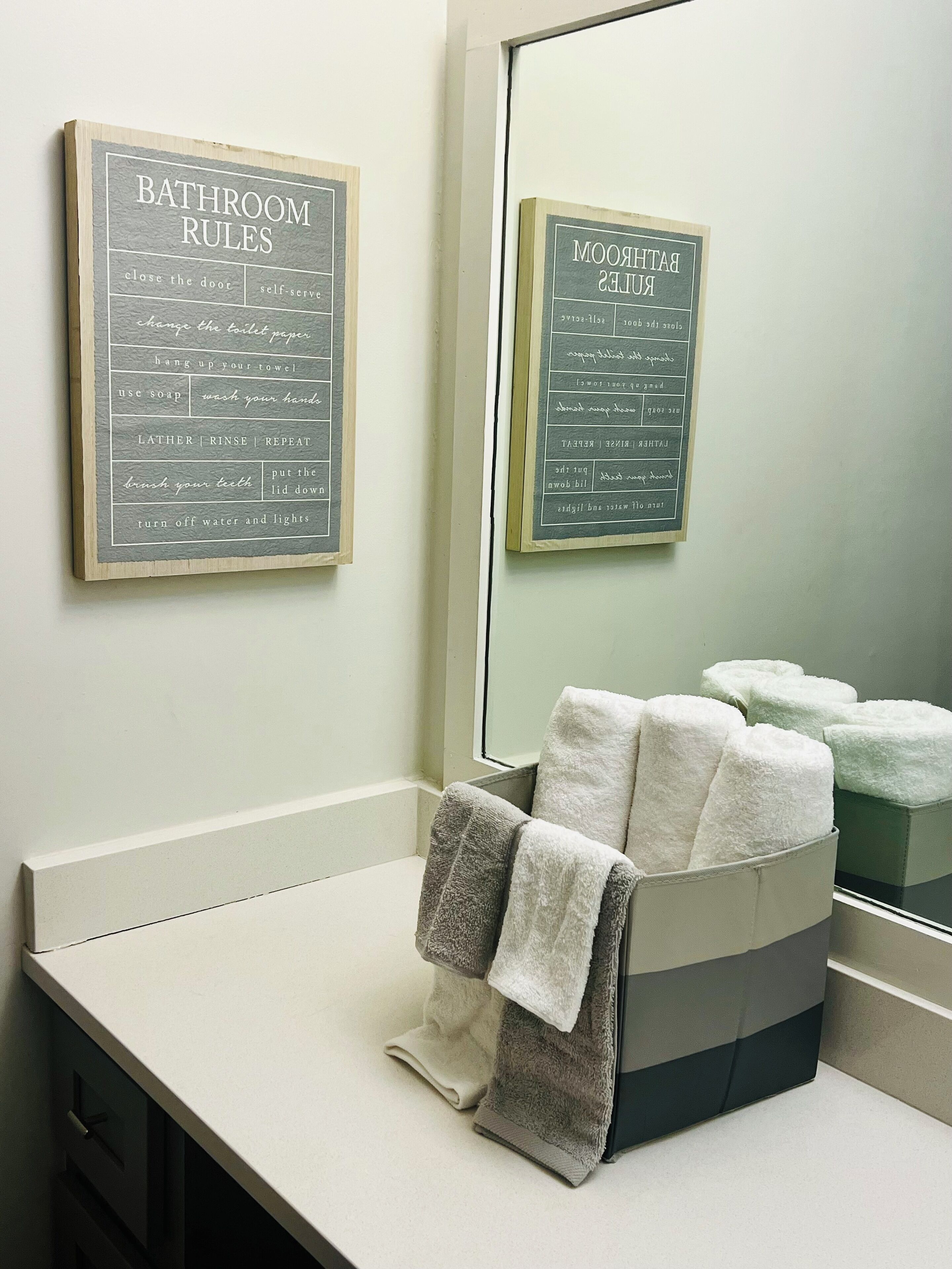 A bathroom with a chalkboard displaying 'Bathroom Rules' and 'Bar Rules'. There are towels on a counter and a mirror reflecting the room.