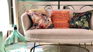 Un canapé beige à la structure métallique, agrémenté de trois coussins à motifs, d’une bouteille en verre et d’un tapis à fleurs.