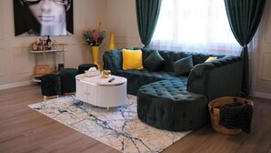 A living room with a sectional sofa, a round coffee table, a side table, a large painting, and a window with curtains. - The Luxe Haven (Cerdanyola del Vallès)
