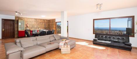 A spacious living room with a large sectional sofa, a smaller sofa, and a black leather couch. There is a dining area with a table and chairs in the background.