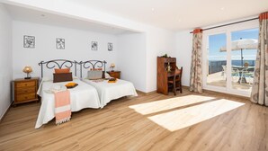 A bedroom with two beds, a wooden desk, a nightstand, and a view of the sea through a sliding glass door.