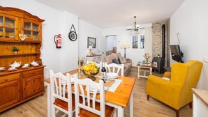 A cozy living room with a wooden dining table, white chairs, a yellow armchair, a fireplace, and a kitchen area with a stove.