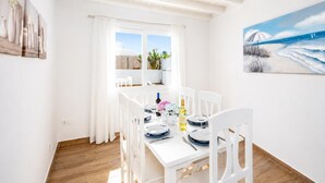 Un comedor con una mesa blanca dispuesta para cuatro personas, sillas blancas, un ventanal con cortinas y un cuadro de una playa en la pared.