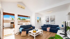 Una sala de estar con dos sofás, una mesa de centro y un televisor. Hay una ventana con vista a una playa y una parrilla afuera.