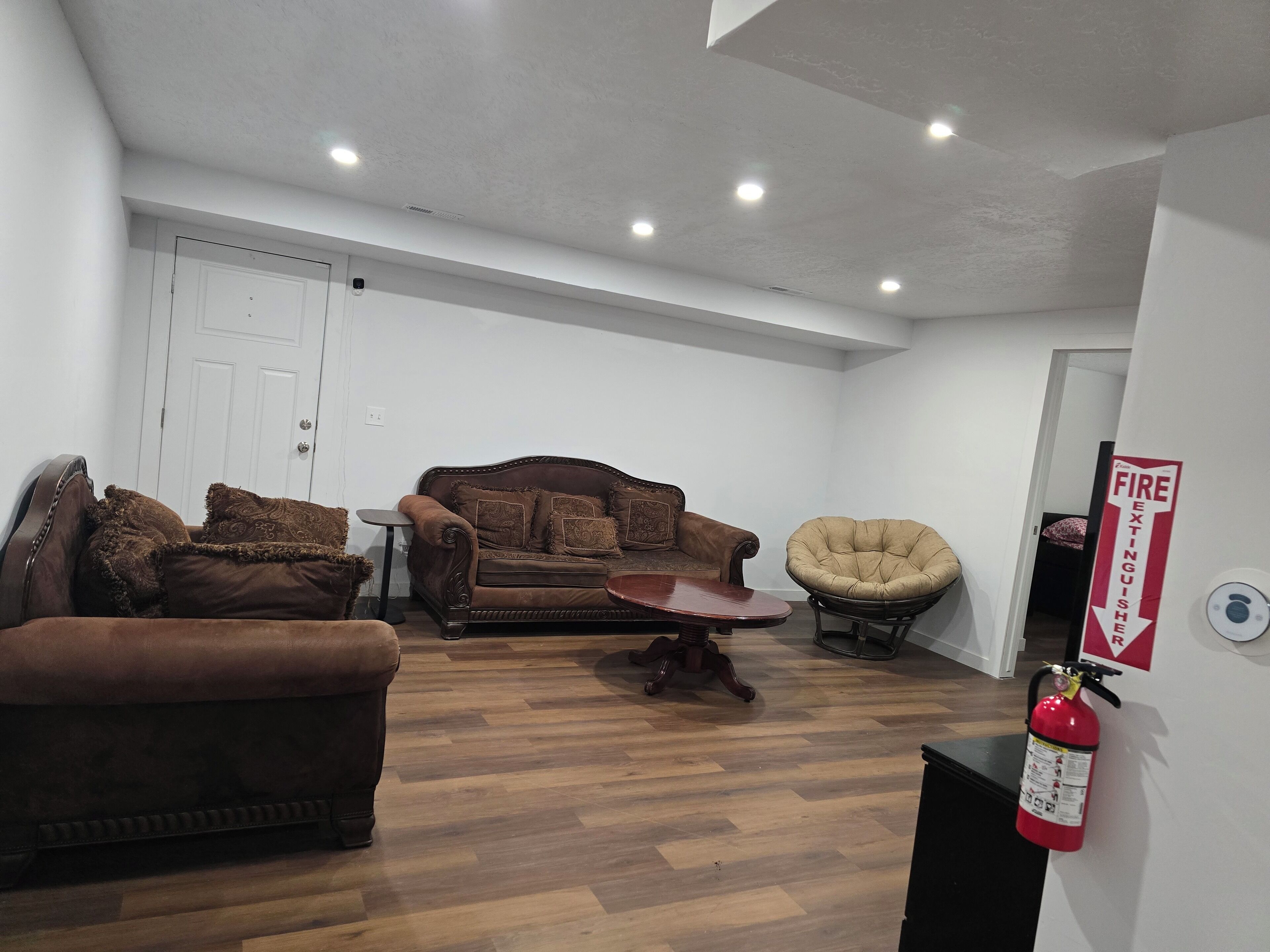 Un salon spacieux avec un canapé en cuir marron, un fauteuil à motifs et une table basse en bois. On peut voir une porte blanche et un extincteur sur le mur.