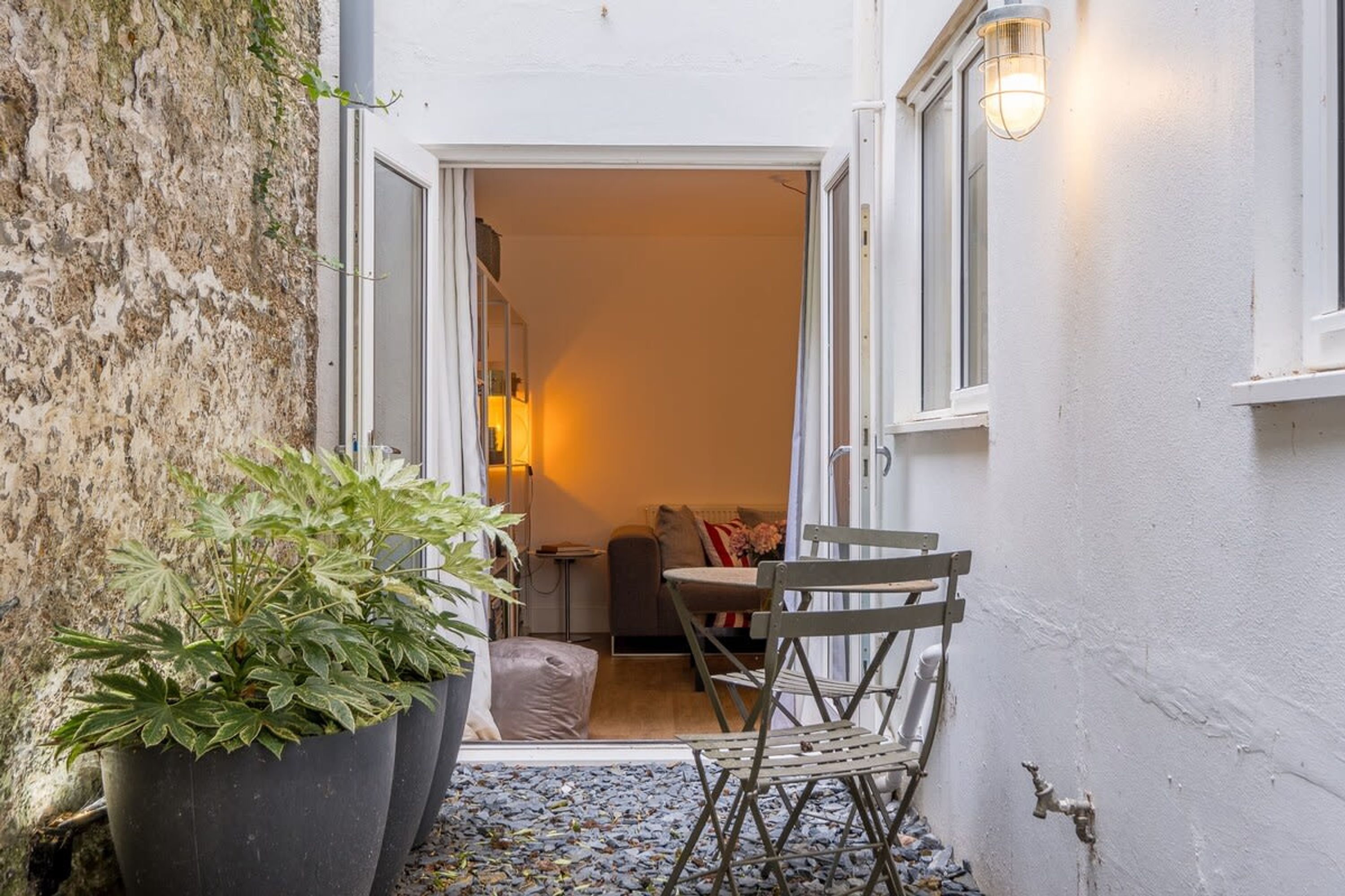 Un petit espace extérieur, agrémenté d’une plante en pot, d’une chaise pliante et d’une porte donnant sur une pièce intérieure où se trouvent un canapé et une lampe.