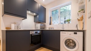 A compact kitchen with dark cabinets, a built-in oven, a washing machine, and a window with a view of greenery. - Host Stay Osbourne Rest (Hove)