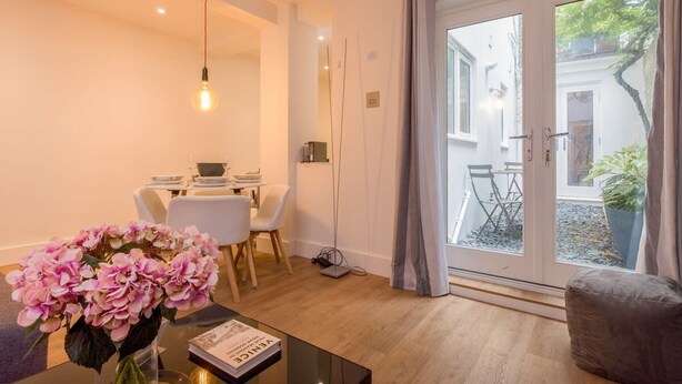 A modern living room with a glass door leading to a garden, a dining area with a table and chairs, and a coffee table with a book and flowers. - Host Stay Osbourne Rest (Hove)