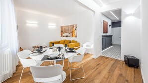 A modern dining area with a wooden floor, white chairs, a yellow sofa, and a dining table set for four. - Emilia Suite Academy (Modena)