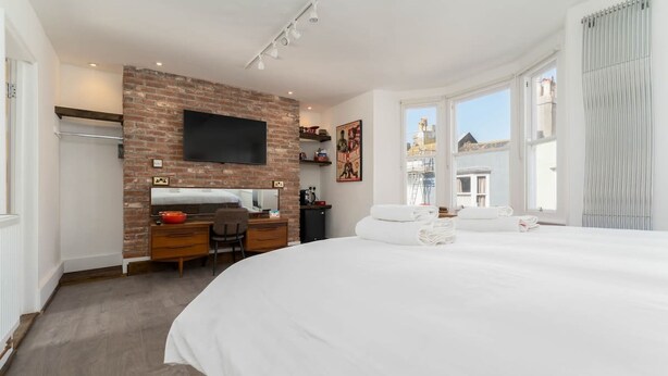 A hotel room with a large bed, a desk with a computer, a flat-screen TV, and a brick accent wall. - Host Stay Grafton Suite (Brighton)