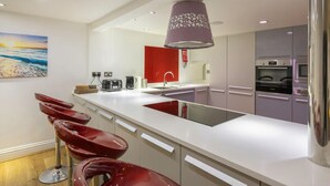 Een moderne keuken met een wit aanrecht, rode barstoelen, een grote hanglamp en roestvrijstalen apparatuur.