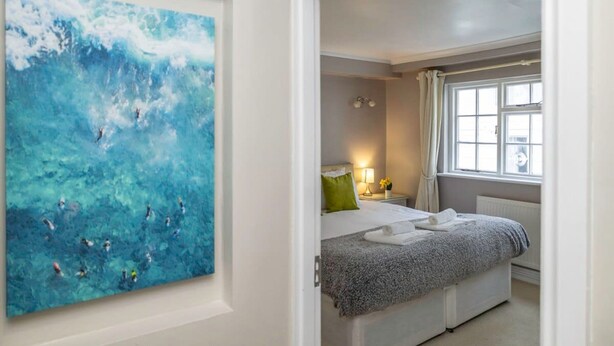 Een slaapkamer met een bed, een nachtkastje, een raam met gordijnen en een schilderij van een blauw zeegezicht.