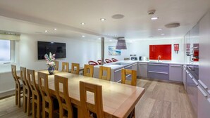 Een moderne keuken met een eethoek, houten stoelen, een grote tafel en een aan de muur bevestigde televisie.
