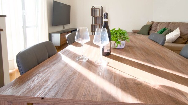 Un tavolo da pranzo con bicchieri e una bottiglia di vino, un divano e una televisione in una stanza ben illuminata.
