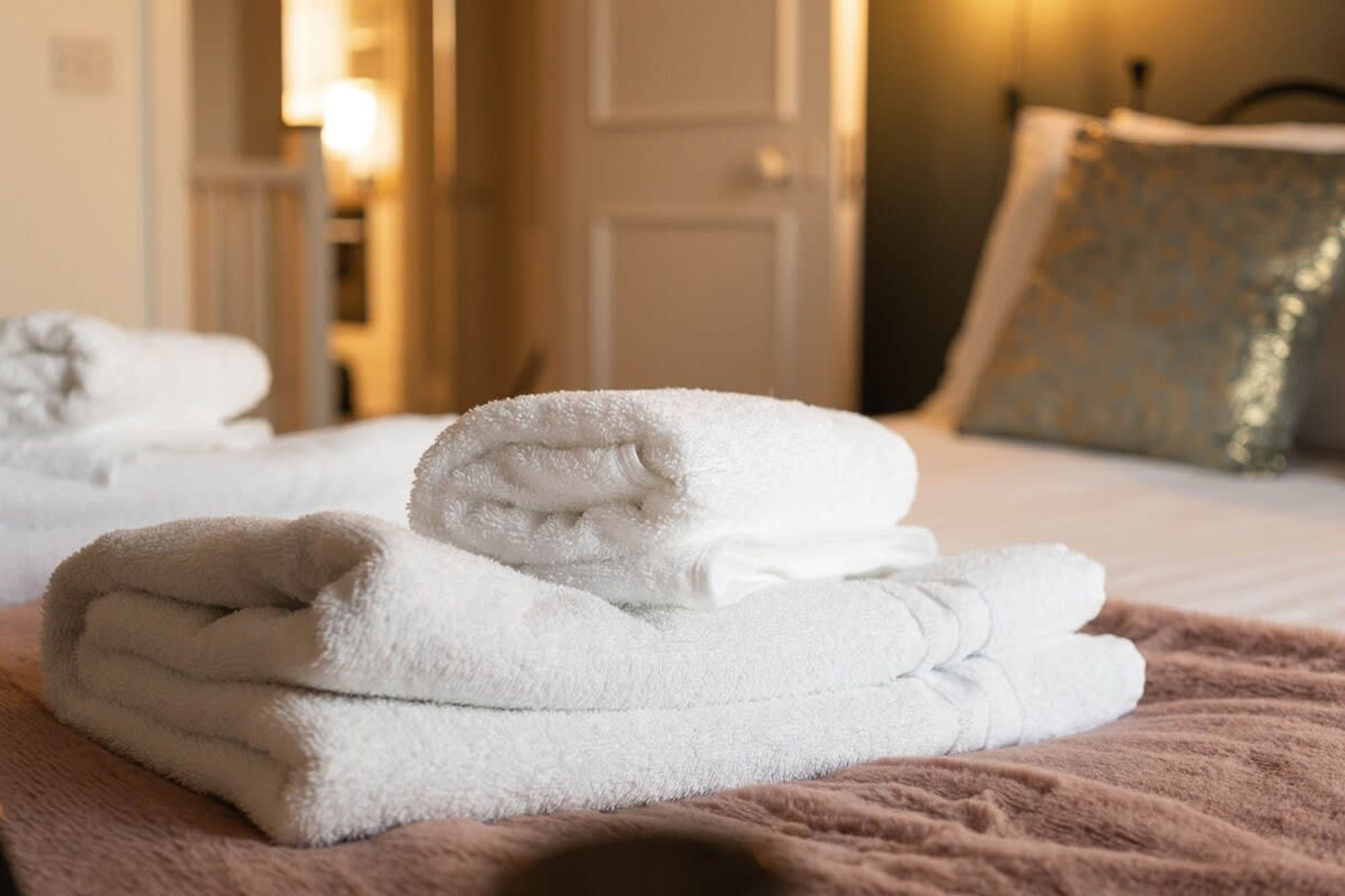 Een keurig opgevouwen witte handdoek op een bed met een bruine deken, een deur op de achtergrond en een kussen met een patroon.