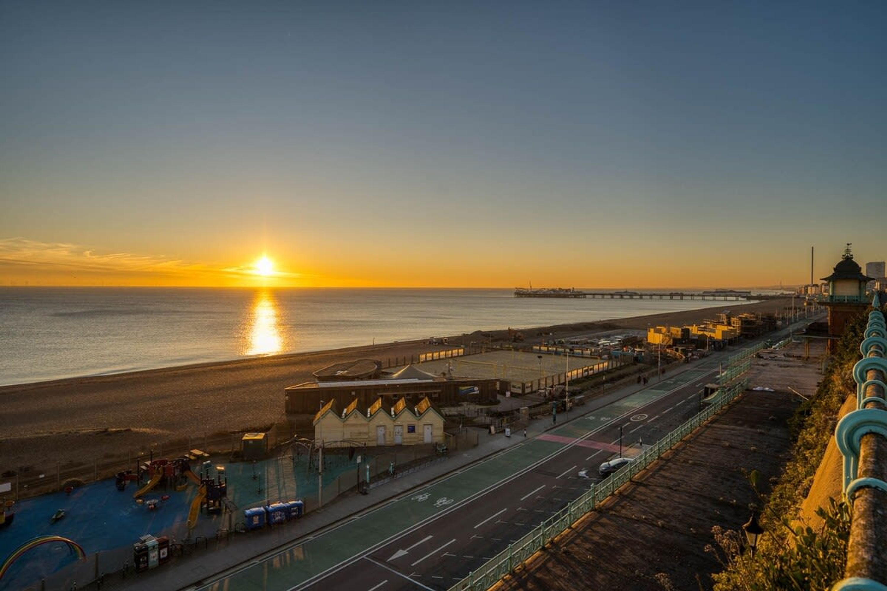 A coastal road with a sunset, a beach, and a bridge in the distance.