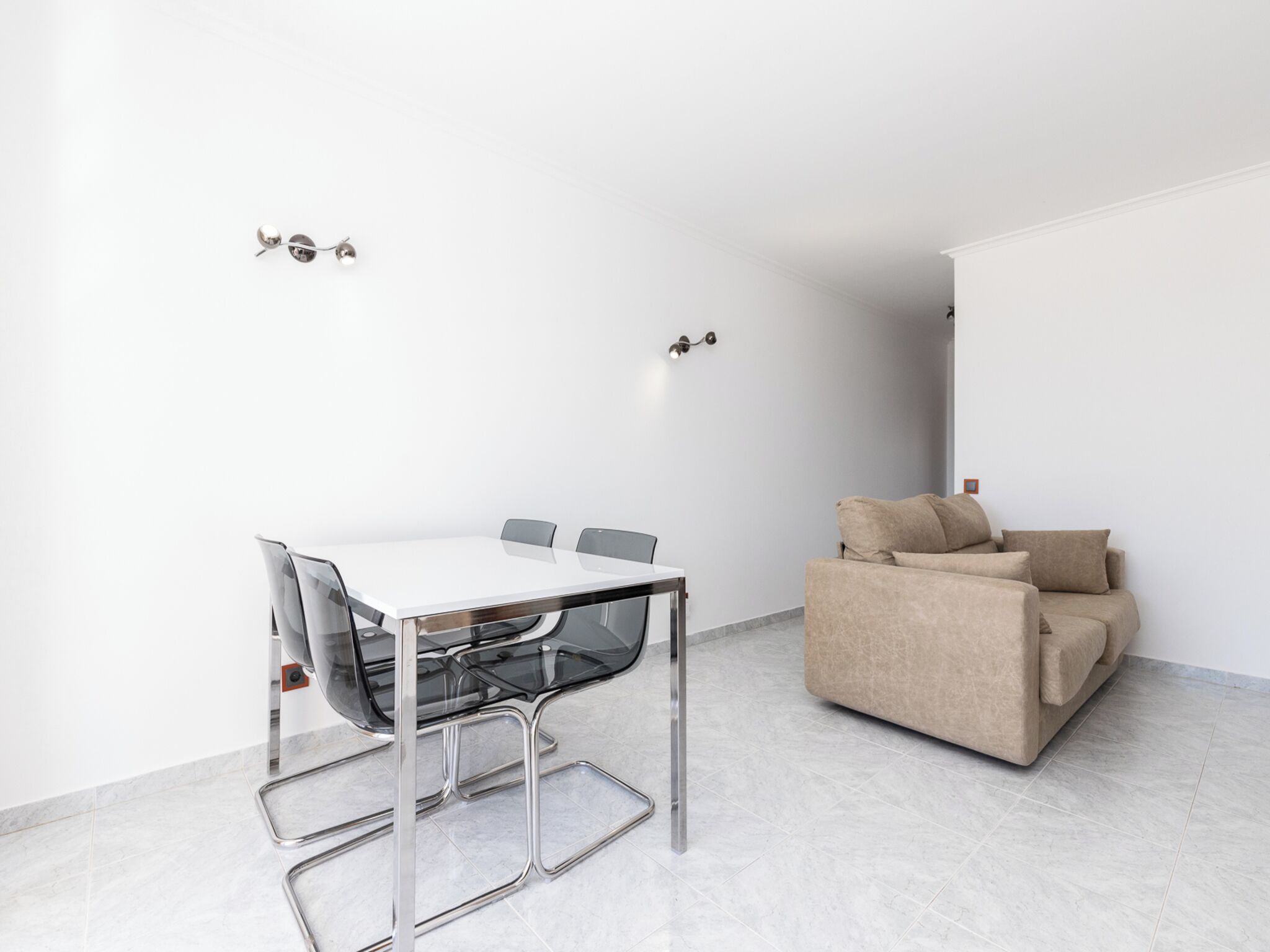 Ein moderner, minimalistischer Raum mit einem weißen Tisch und Stühlen, einem beigen Sofa und einem Fliesenboden.