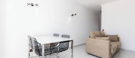 Ein moderner, minimalistischer Raum mit einem weißen Tisch und Stühlen, einem beigen Sofa und einem Fliesenboden.