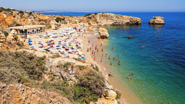 A beach with a building on the left, clear blue water, and rocky terrain. - Ory Red Apartment, São Rafael, Albufeira (Albufeira)