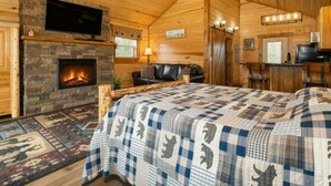 A cozy log cabin interior with a fireplace, a large bed, a television, and a dining area.