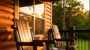 A wooden rocking chair and a table on a porch with a view of trees and a window.
