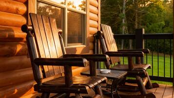 A wooden rocking chair and a table on a porch with a view of trees and a window.