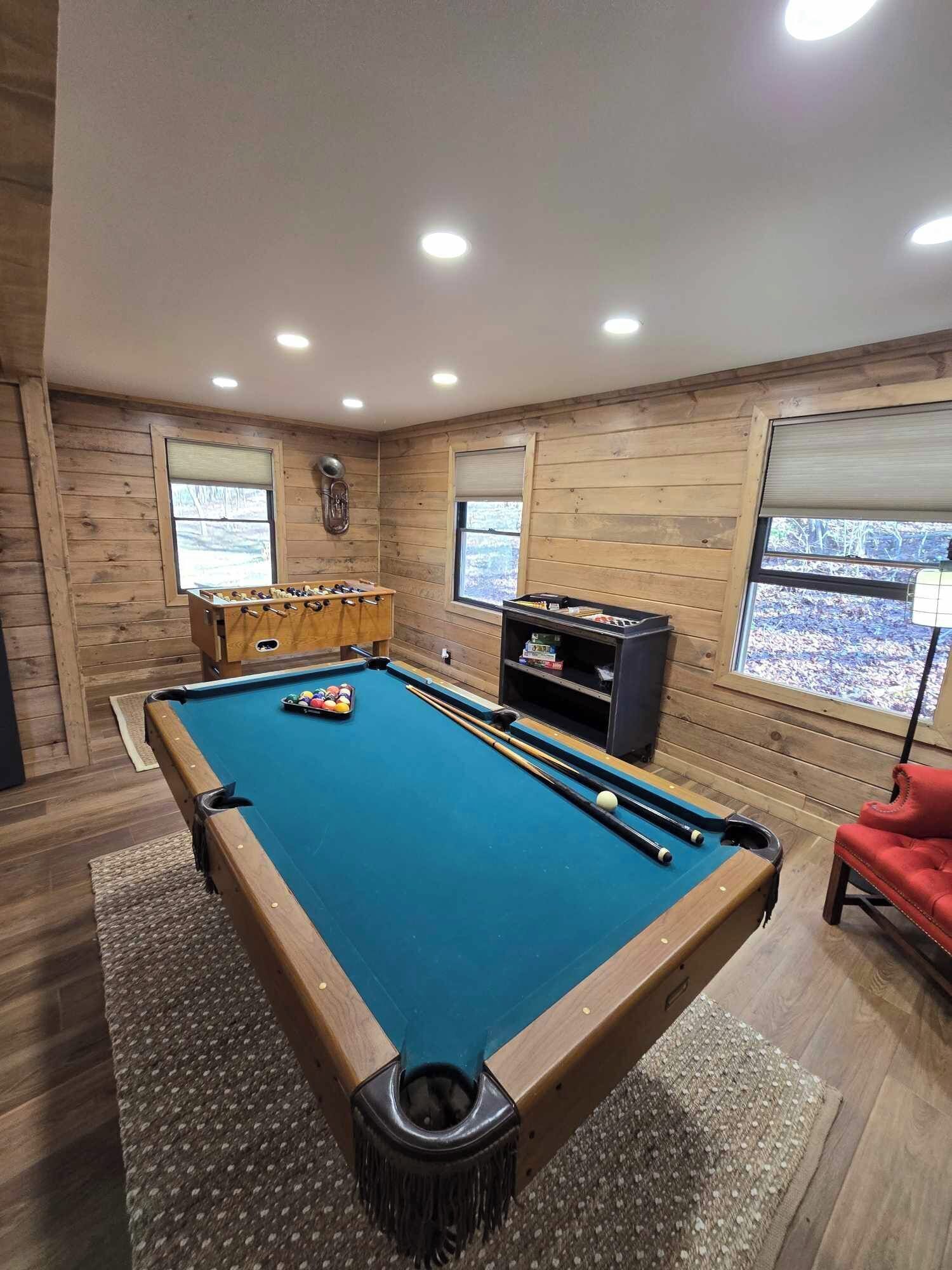 Een pooltafel, een tafelvoetbalspel en een biljarttafel in een ruimte met houten wanden en grote ramen.