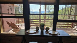 Una mesa de madera con dos sillas, un ventanal con vista a una terraza de madera y árboles, y dos tazas con huellas de patas.