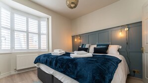 Quarto com uma cama grande, roupa de cama branca, cabeceira azul escura e uma janela com persianas.