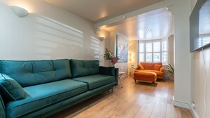 Uma sala de estar com um sofá verde, uma poltrona laranja, piso de madeira, uma TV de tela plana na parede e uma janela com persianas.