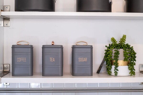 A kitchen shelf with three grey containers labeled 'Coffee,' 'Tea,' and 'Sugar,' and a small potted plant.