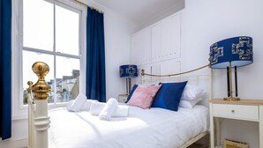 A neatly made bed with white linens, a bedside table with a lamp, and a window with blue curtains. - Host Stay Guildford Apartment (Brighton)