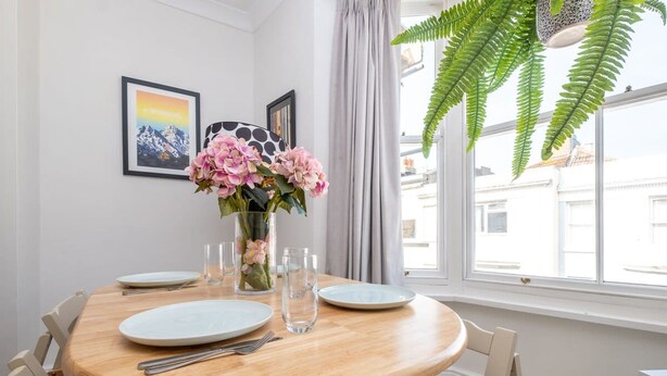 A dining area with a round wooden table, white plates, cutlery, and a vase with pink flowers. There are framed pictures on the wall and a large window with a view of buildings outside. - Host Stay Guildford Apartment (Brighton)
